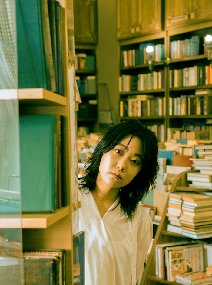 A woman in a white shirt looks contemplatively from behind a bookshelf in a cozy, well-stocked bookstore.